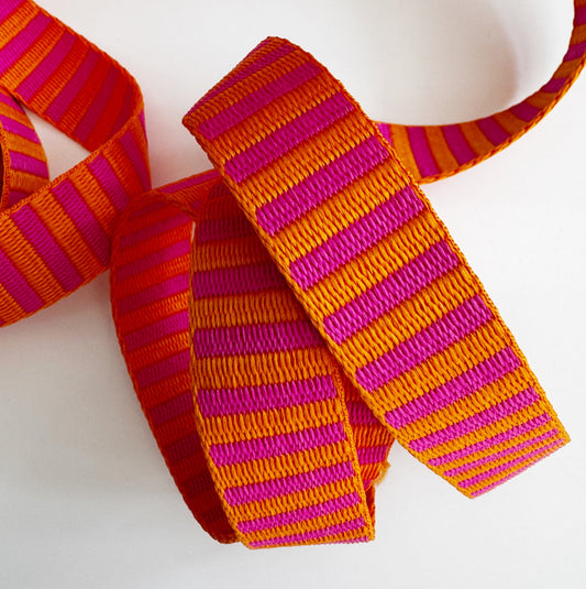 Closed up shot of 1.5-inch wide jacquard woven webbing strap showcasing a vibrant hot pink and orange horizontal stripe pattern, loosely coiled against a white background.
