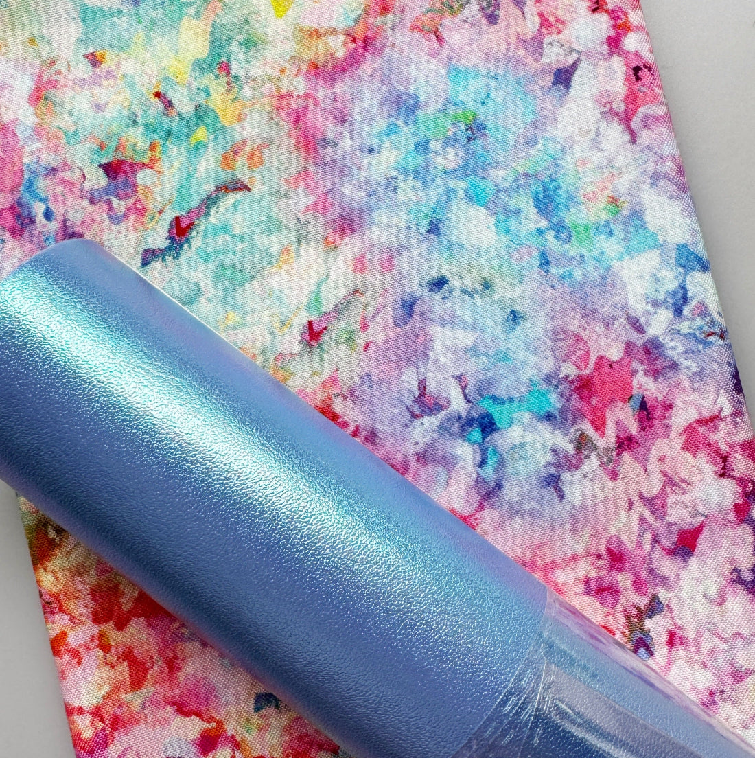 A close-up of a rolled sheet of iridescent light blue vinyl or craft film placed diagonally over a colorful abstract-patterned sheet. The main product appears to be the blue iridescent material rather than the background patterned sheet.