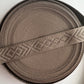 A close-up of a rolled woven strap or trim in a taupe-brown color, featuring a repeating geometric diamond pattern on the center band.