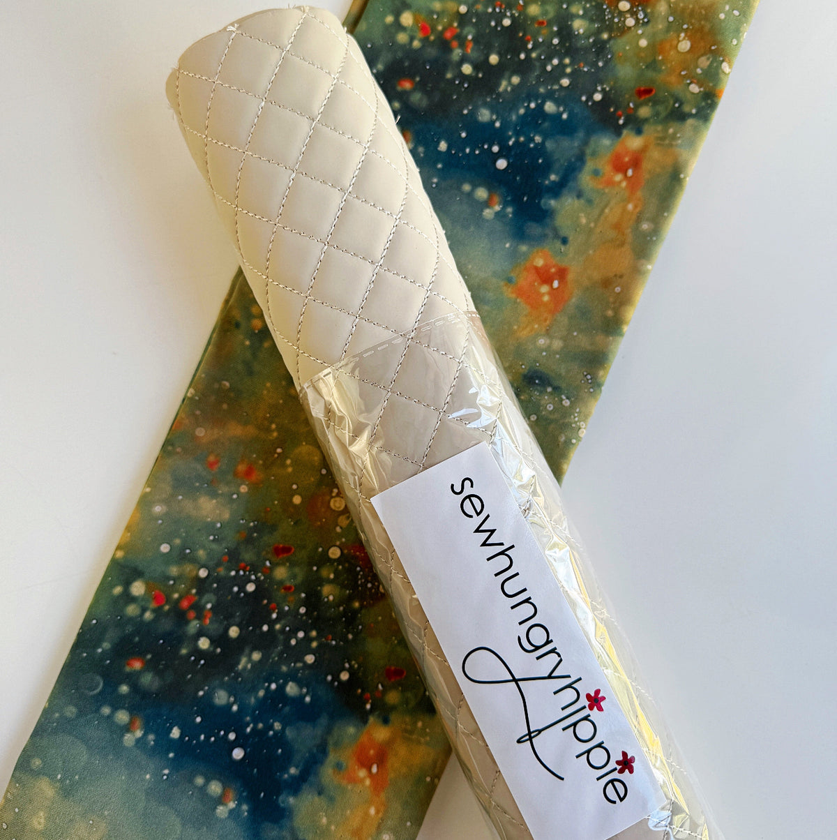 A roll of quilted off-white or cream faux leather vinyl fabric labeled with the SewHungryhippie brand, paired with a colorful, abstract printed fabric background