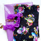DIY bag-making materials featuring Windham Fabrics Serafina Songbirds Black floral cotton fabric by Whistler Studios, coordinating solid purple fabric, and a purple acrylic chain purse strap with gold-tone lobster clasps. A colorful fabric and hardware combination for sewing custom handbags, shoulder bags, and handmade accessories.