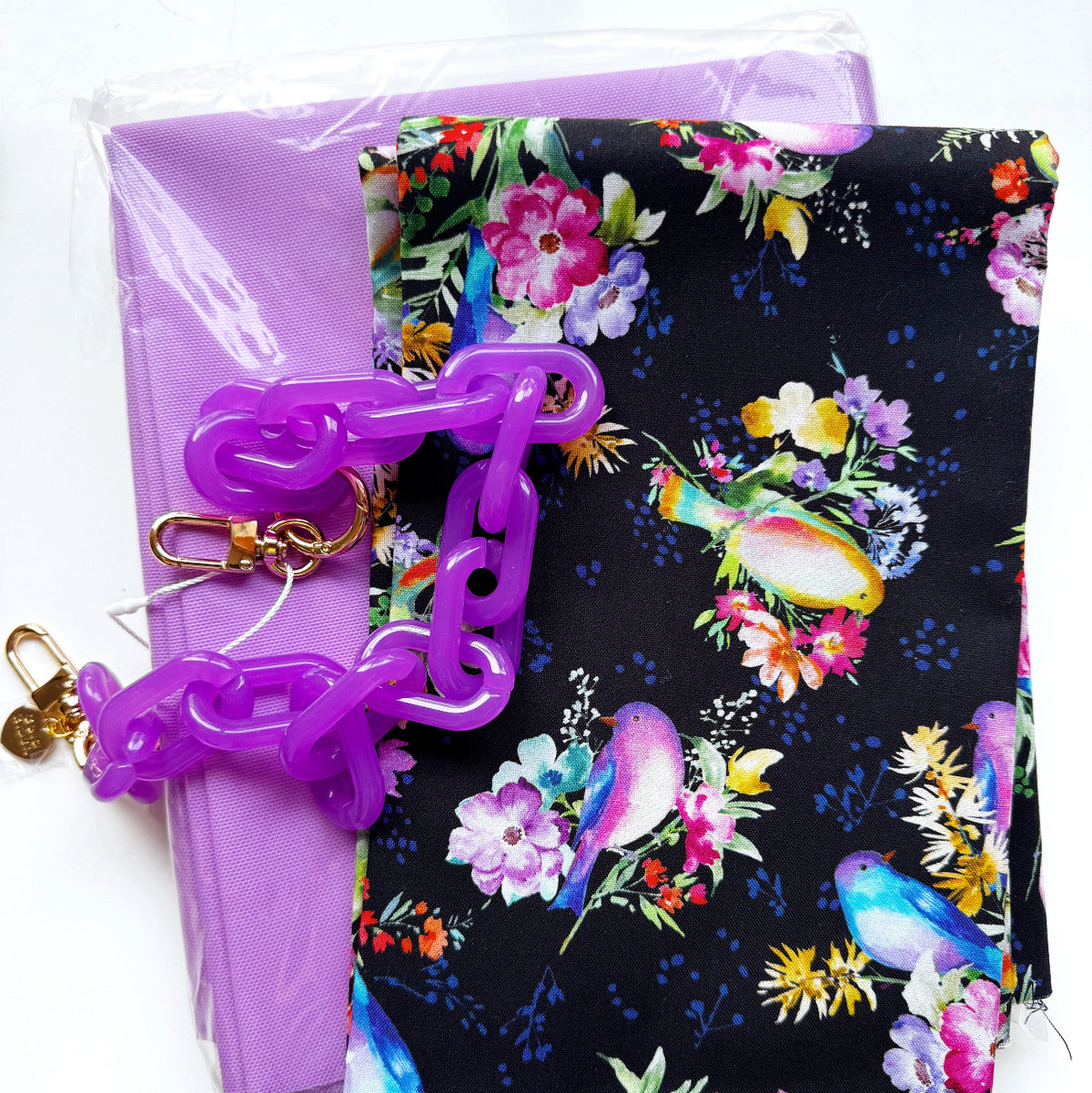 DIY bag-making materials featuring Windham Fabrics Serafina Songbirds Black floral cotton fabric by Whistler Studios, coordinating solid purple fabric, and a purple acrylic chain purse strap with gold-tone lobster clasps. A colorful fabric and hardware combination for sewing custom handbags, shoulder bags, and handmade accessories.
