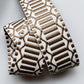 A close-up of a woven elastic strap or trim featuring a geometric pattern in brown and off-white, folded neatly on a light gray background.