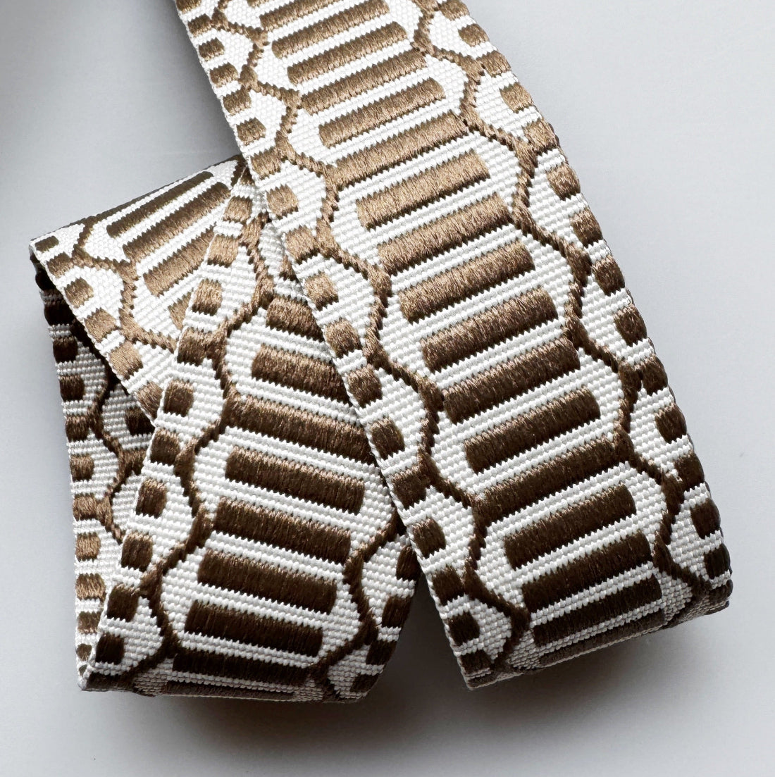 A close-up of a woven elastic strap or trim featuring a geometric pattern in brown and off-white, folded neatly on a light gray background.