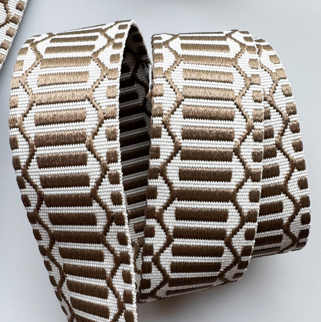 A close-up of a woven ribbon or trim with a geometric pattern in brown and off-white, rolled into loops on a light gray background.