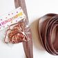 A brown woven strap with a rose gold metal clasp, shown next to its clear plastic packaging with a white paper label that reads 'HUNGRYHIPPIE'. The strap is arranged on a clean white background.