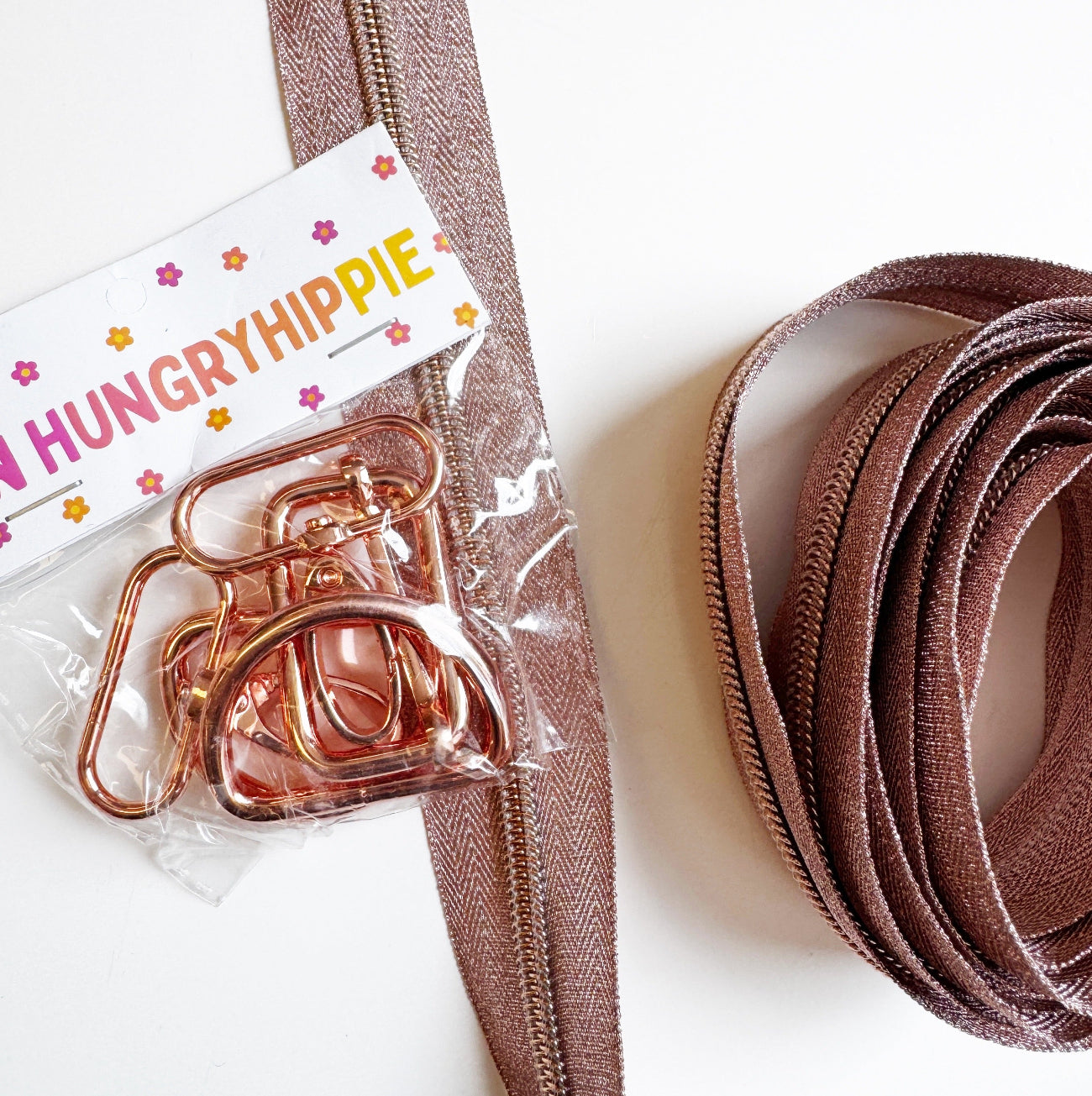 A brown woven strap with a rose gold metal clasp, shown next to its clear plastic packaging with a white paper label that reads 'HUNGRYHIPPIE'. The strap is arranged on a clean white background.