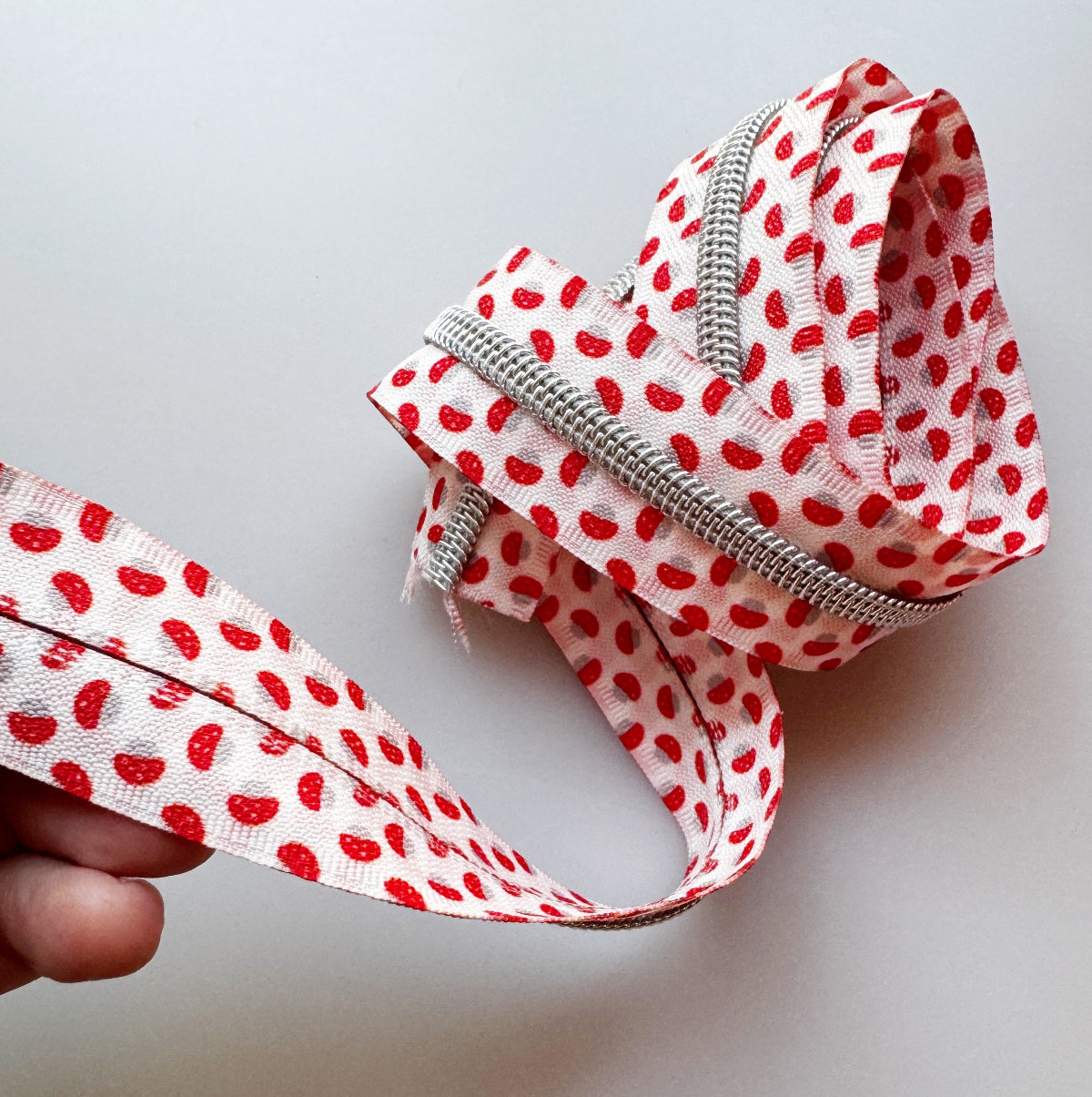 A close-up of a silver metal zipper with a white fabric tape printed with a red strawberry pattern, held by a hand against a plain light gray background.