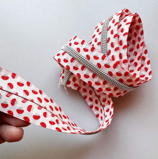 Hand holding a loop of white #5 zipper tape printed with red mushroom caps and silver coil teeth
