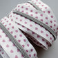 A close-up of a white zipper tape featuring a pattern of small pink hearts, with a silver metal zipper running along the edge, set against a neutral gray background.