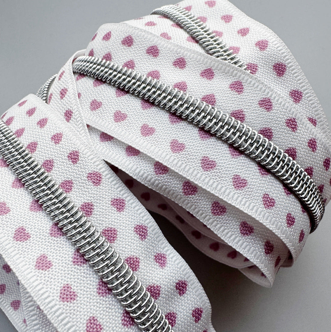 A close-up of a white zipper tape featuring a pattern of small pink hearts, with a silver metal zipper running along the edge, set against a neutral gray background.