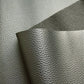 A close-up view of textured, dark grey faux leather or vinyl fabric draped in soft folds against a plain white background. The material features a fine, pebbled grain surface that subtly reflects the light.
