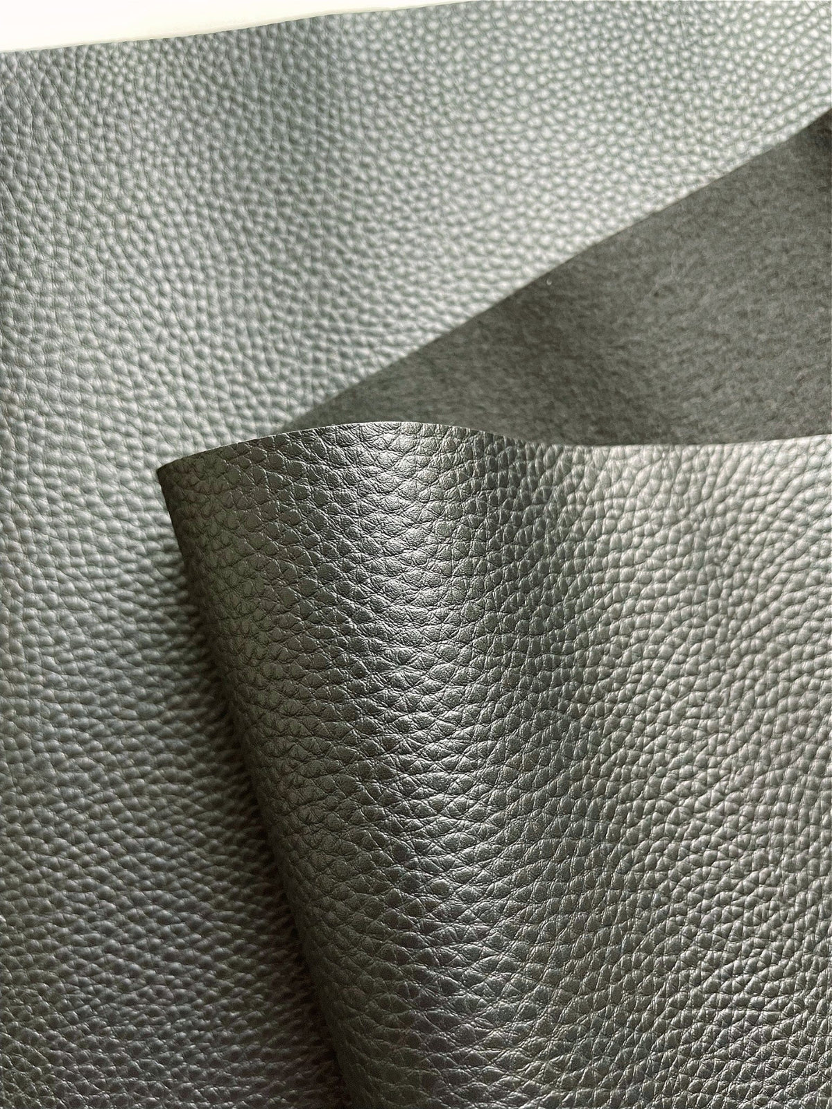 A close-up view of textured, dark grey faux leather or vinyl fabric draped in soft folds against a plain white background. The material features a fine, pebbled grain surface that subtly reflects the light.