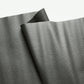 A close-up view of textured, dark grey faux leather or vinyl fabric draped in soft folds against a plain white background. The material features a fine, pebbled grain surface that subtly reflects the light.