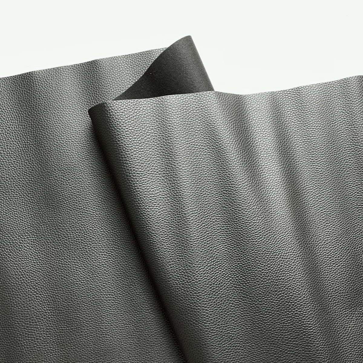 A close-up view of textured, dark grey faux leather or vinyl fabric draped in soft folds against a plain white background. The material features a fine, pebbled grain surface that subtly reflects the light.
