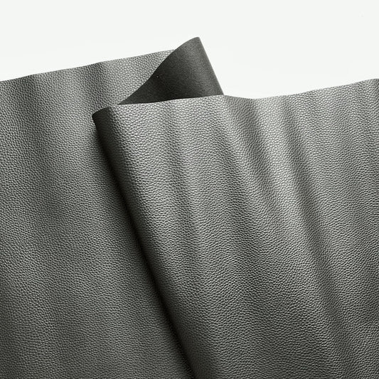 A close-up view of textured, dark grey faux leather or vinyl fabric draped in soft folds against a plain white background. The material features a fine, pebbled grain surface that subtly reflects the light.